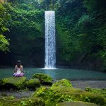 A woman wearing pink purplish colour is sitting cross-legged on a rock in front of a single drop waterfall on a moss-covered cove, with her hands placed on her knee. Tibumana Waterfall. Bali waterfalls tour | Bespoke Indonesia Holiday.
