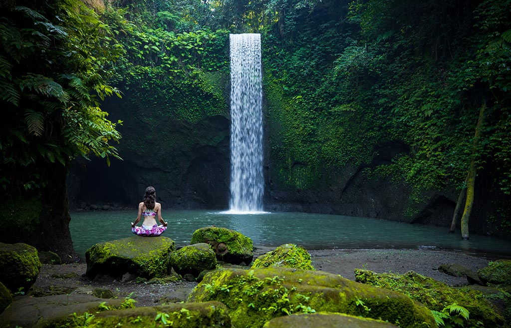 A woman wearing pink purplish colour is sitting cross-legged on a rock in front of a single drop waterfall on a moss-covered cove, with her hands placed on her knee. Tibumana Waterfall. Bali waterfalls tour | Bespoke Indonesia Holiday.