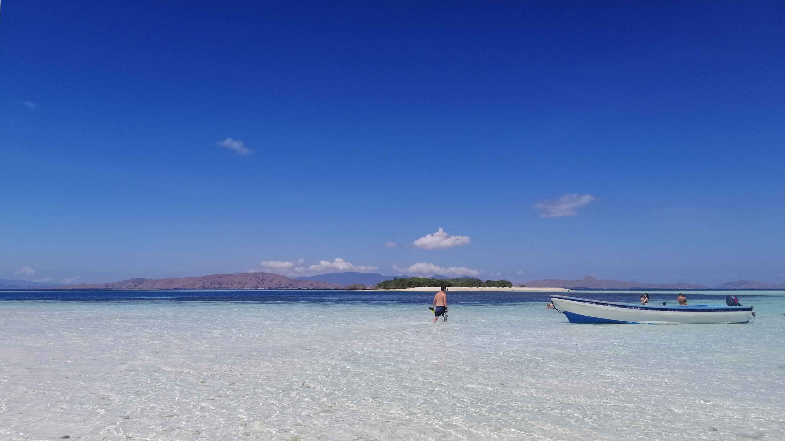 In the middle of the picture, a man is wading in the clear beach water. On the right side, a white small boat. The sky is bright blue. Manta Point | Komodo Leisure Cruise | Bespoke Indonesia Holiday.