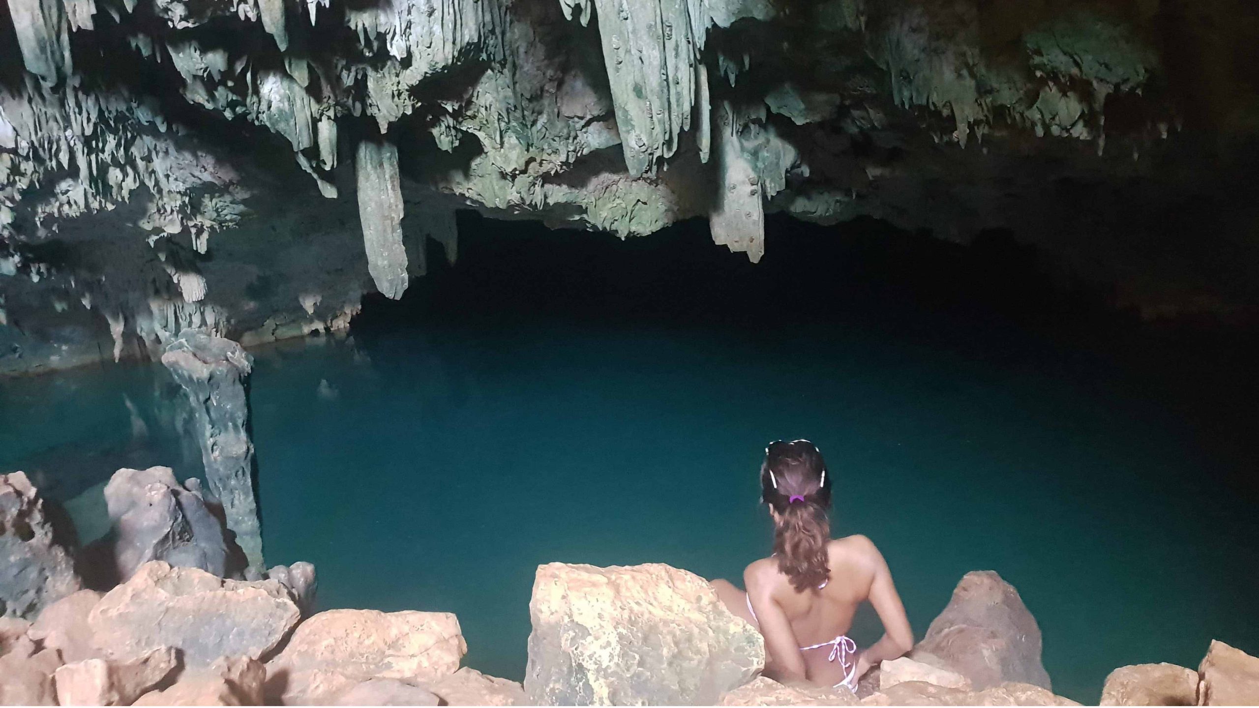 A view of a stalactite cave with water. The girl is sitting in a quarry and is going for a swim. Rangko Cave | Bespoke Indonesia Holiday.