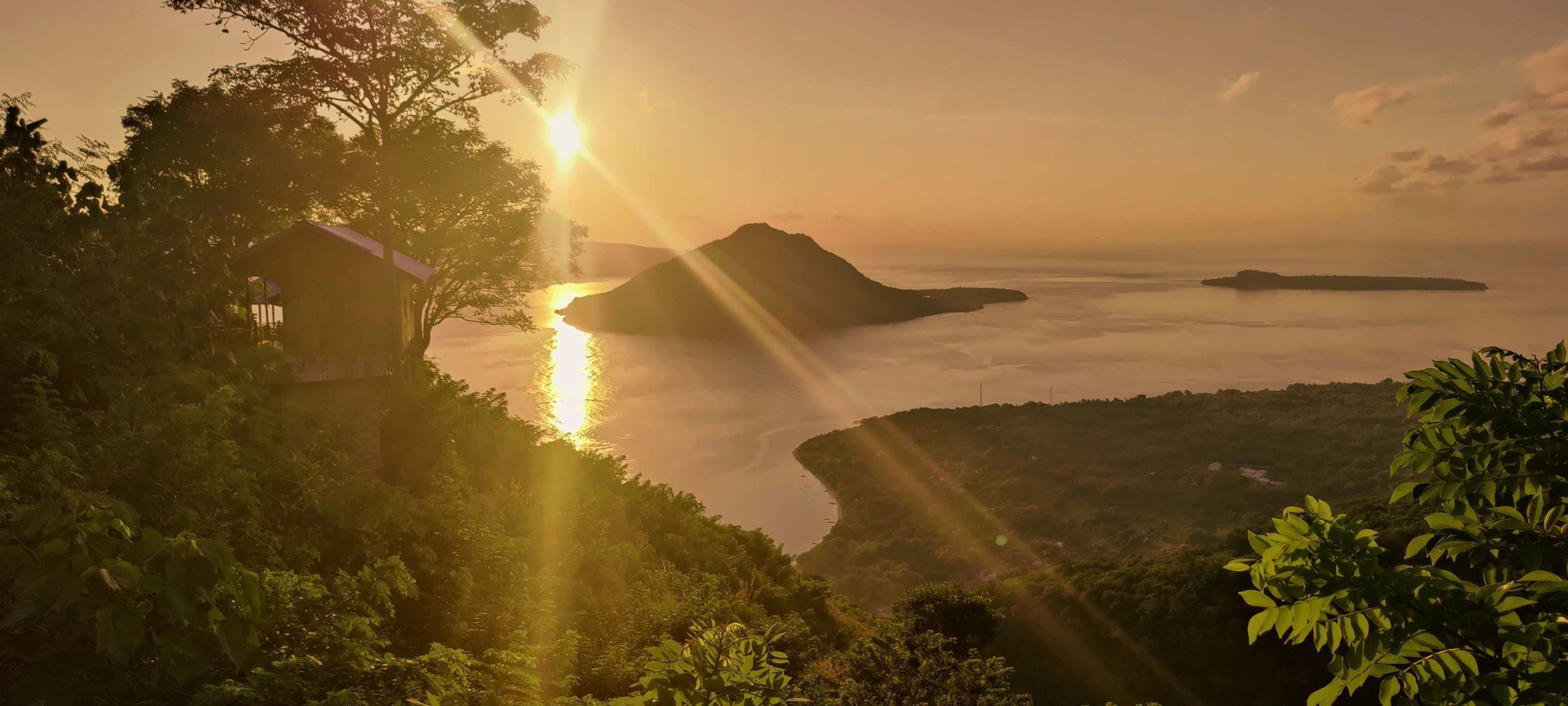 Sunset view from a high hill to the sea. There is a small cabin on the left side of the hill. There is a high island in the sea behind which the sun sets. Where to Go in Indonesia?| Tailor Made Holidays Indonesia.