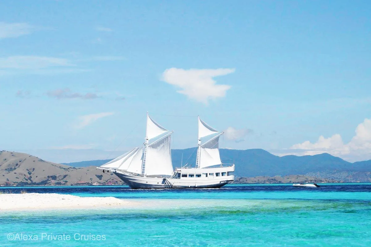 A beautiful white boat with hills in the background and white sand bar in the foreground. Alexa Yacht. Alexa private cruises. Indonesia yacht charter. Luxury cruises Indonesia. Bespoke Indonesia Holiday