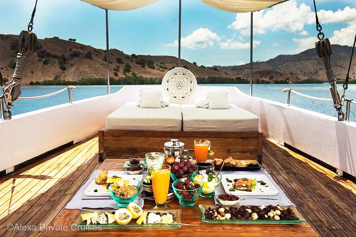 A table full of food and two glasses of juice positioned in front of a large sun lounger on a aft deck of a wooden sailing boat. Alexa private cruises. Alexa yacht. Luxury Cruise Indonesia. Yacht charter Indonesia. Honeymoon cruise in Indonesia. Bespoke Indonesia Holiday.