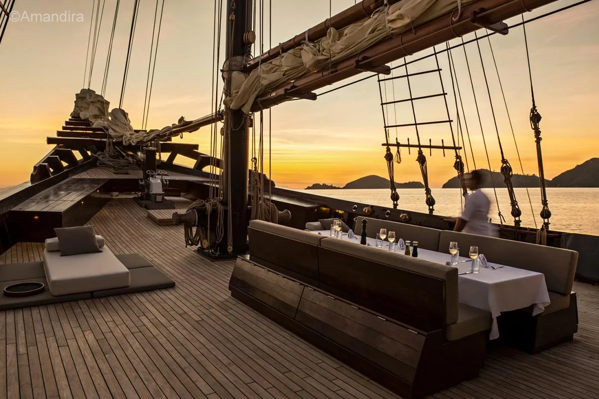 A guy in a white top is passing a dining table setup on an open deck on a wooden sailing boat. Amandira. Luxury holidays Indonesia. Luxury Cruise Indonesia. Yacht charter Indonesia. Private cruise Indonesia. Sailing trip Indonesia. Yacht Rental in Indonesia. Bespoke Indonesia Holiday.