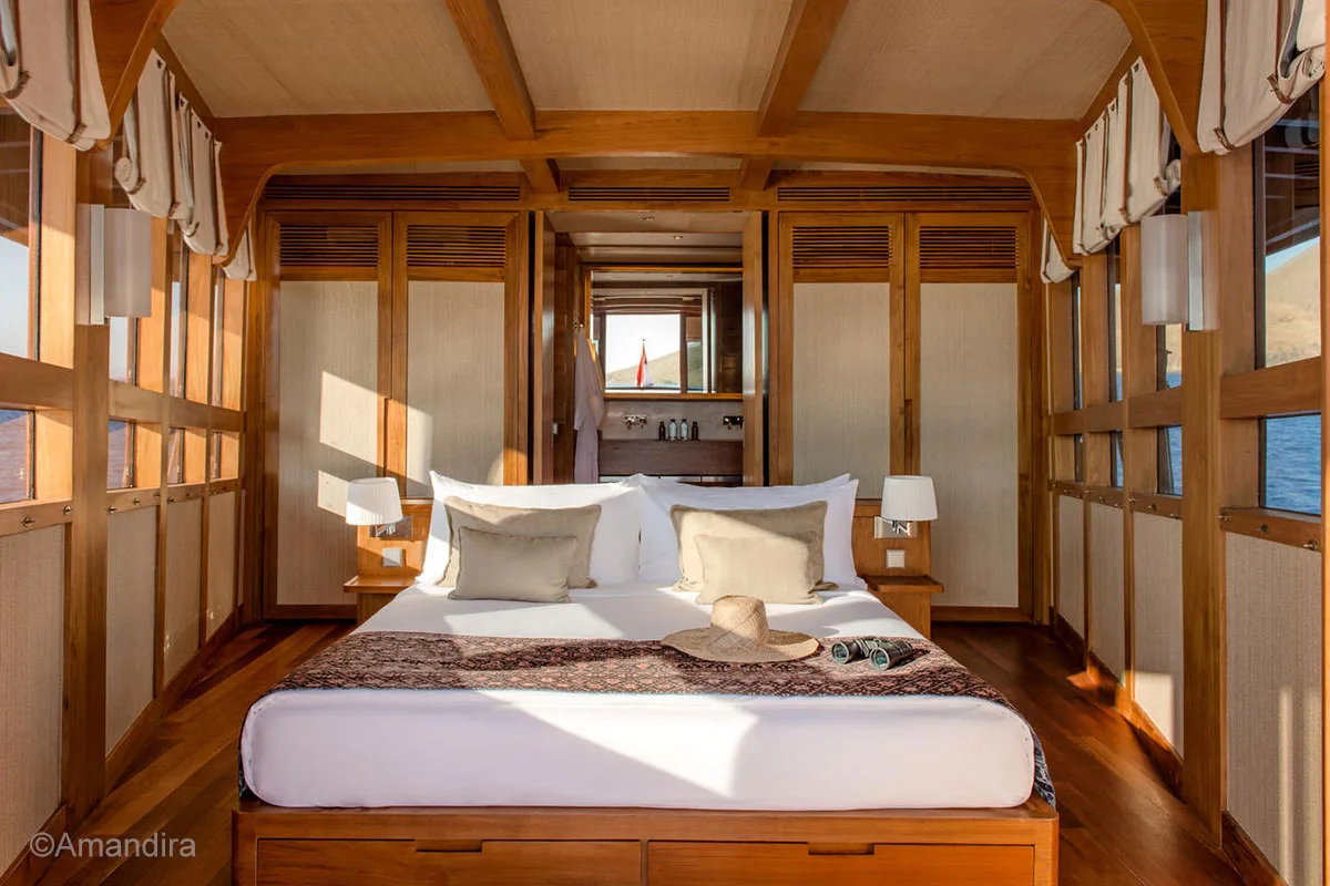 A king-sized bed surrounded by windows and there is a washbasin behind the window separated by wooden room dividers. Amandira. Aman Yacht. Luxury Cruise Indonesia. Yacht charter Indonesia. Indonesia private cruise. luxury boat rental Indonesia. Luxury Indonesia holiday. Bespoke Indonesia Holiday.
