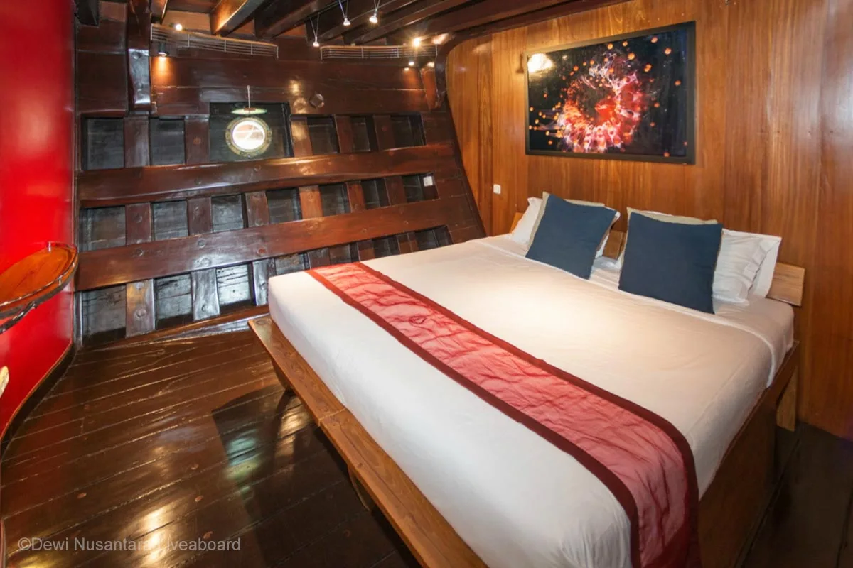 A wooden with a king-sized bed adorned with navy blue cushions and red orangish runner, and a painting above the headboard. Dewi Nusantara Liveaboard. Liveaboard Indonesia. Luxury liveaboard Indonesia. Luxury liveaboard Raja Ampat. Luxury liveaboard Komodo. Diving expeditions Indonesia. Liveaboard tours Indonesia. Diving trip Indonesia. Private liveaboard trip Indonesia. Bespoke Indonesia Holiday.
