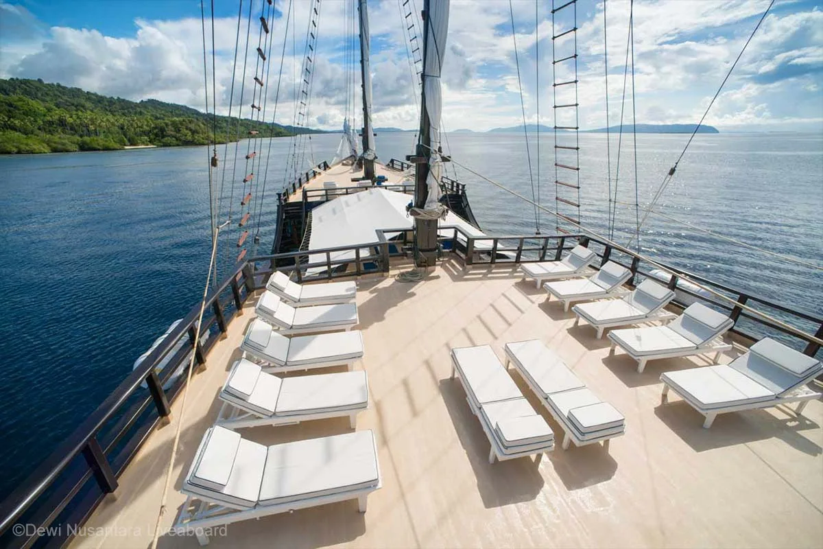 There are 12 white loungers on the top of a sailing boat that is cruising by a green lush island on the side. Dewi Nusantara Liveaboard. Liveaboard Indonesia. Luxury liveaboard Indonesia. Luxury liveaboard Raja Ampat. Luxury liveaboard Komodo. Diving expeditions Indonesia. Liveaboard tours Indonesia. Private liveaboard trip Indonesia. Bespoke Indonesia Holiday.