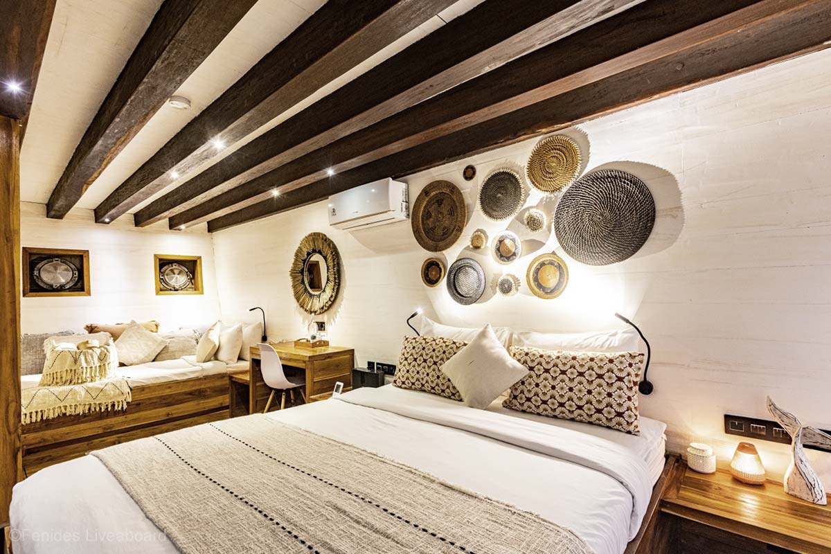 Wooden bedroom predominantly by white and natural brown colour, featuring queen-size bed, bedside tables, working table, and a wooden couch. Fenides. Fenides Liveaboard. Liveaboard Indonesia. Luxury liveaboard Indonesia. Luxury liveaboard Raja Ampat. Luxury liveaboard Komodo. Surfing trip Indonesia. Diving expeditions Indonesia. Indonesia Diving trip. Private liveaboard trip Indonesia. Luxury holidays Indonesia. Luxury Cruise Indonesia. Bespoke cruise Indonesia. Yacht charter Indonesia. Yacht charter Komodo. Yacht charter Raja Ampat. Private cruise Indonesia. Sailing trip Indonesia. Yacht Rental in Indonesia. Bespoke Indonesia Holiday.