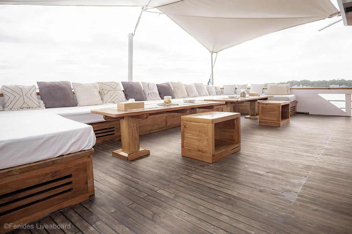 Large wooden lounge with white mattress, two long wooden tables, and two square wooden ottomans on a wooden boat under a white shade. Fenides. Fenides Liveaboard. Liveaboard Indonesia. Luxury liveaboard Indonesia. Luxury liveaboard Raja Ampat. Luxury liveaboard Komodo. Surfing trip Indonesia. Diving expeditions Indonesia. Indonesia Diving trip. Private liveaboard trip Indonesia. Luxury holidays Indonesia. Luxury Cruise Indonesia. Bespoke cruise Indonesia. Yacht charter Indonesia. Yacht charter Komodo. Yacht charter Raja Ampat. Private cruise Indonesia. Sailing trip Indonesia. Yacht Rental in Indonesia. Bespoke Indonesia Holiday.