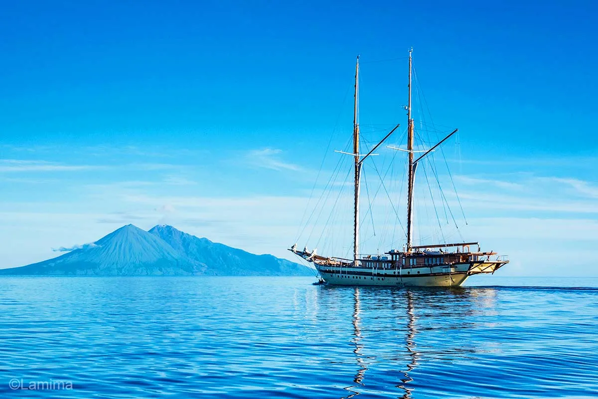 A wooden sailing boat sailing on a blue sea on the right heading to a two triangles volcanoes on the left. Lamima. Lamima yacht. Sailing boat. Luxury Cruise Indonesia. Yacht charter Indonesia. Indonesia private cruise. Tailor made holiday Indonesia. Bespoke Indonesia Holiday.