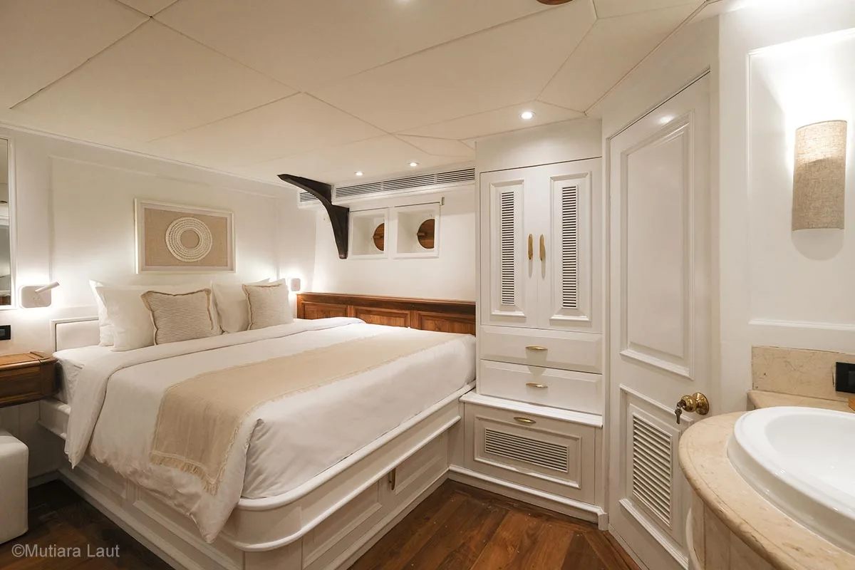 A beautiful bedroom predominantly by white colour with a small portion of brown colour on the floor, dressing table, and the panel on the right side of the queen-sized bed. Mutiara Laut Liveaboard. Liveaboard Indonesia. Luxury liveaboard Indonesia. Luxury liveaboard Raja Ampat. Luxury liveaboard Komodo. Diving expeditions Indonesia. Indonesia Diving trip. Private liveaboard trip Indonesia. Luxury holidays Indonesia. Luxury Cruise Indonesia. Bespoke cruise Indonesia. Yacht charter Indonesia. Yacht charter Komodo. Yacht charter Raja Ampat. Private cruise Indonesia. Sailing trip Indonesia. Yacht Rental in Indonesia. Bespoke Indonesia Holiday.