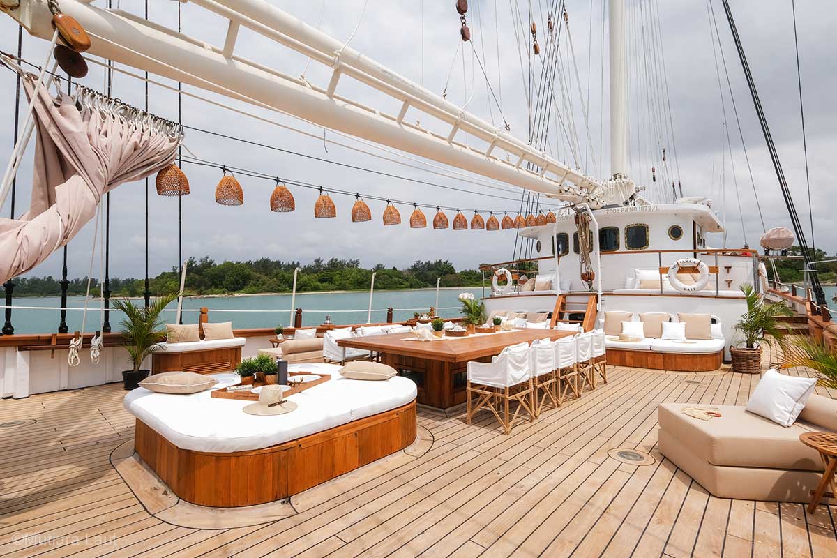 A spacious deck with many rattan lamp shades and curtain hanging on the rope under the mast, alfresco dining, lounge seating, and a large sun lounger. Fenides. Fenides Liveaboard. Liveaboard Indonesia. Luxury liveaboard Indonesia. Luxury liveaboard Raja Ampat. Luxury liveaboard Komodo. Surfing trip Indonesia. Diving expeditions Indonesia. Indonesia Diving trip. Private liveaboard trip Indonesia. Luxury holidays Indonesia. Luxury Cruise Indonesia. Bespoke cruise Indonesia. Yacht charter Indonesia. Yacht charter Komodo. Yacht charter Raja Ampat. Private cruise Indonesia. Sailing trip Indonesia. Yacht Rental in Indonesia. Bespoke Indonesia Holiday.