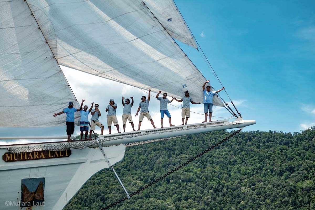 Nine men in light blue uniform are standing on a vertical white pole at the edge of a white wooden boat under the white sails. Mutiara Laut crew members. Mutiara Laut Liveaboard. Liveaboard Indonesia. Luxury liveaboard Indonesia. Luxury liveaboard Raja Ampat. Luxury liveaboard Komodo. Diving expeditions Indonesia. Indonesia Diving trip. Private liveaboard trip Indonesia. Luxury holidays Indonesia. Luxury Cruise Indonesia. Bespoke cruise Indonesia. Yacht charter Indonesia. Yacht charter Komodo. Yacht charter Raja Ampat. Private cruise Indonesia. Sailing trip Indonesia. Yacht Rental in Indonesia. Bespoke Indonesia Holiday.