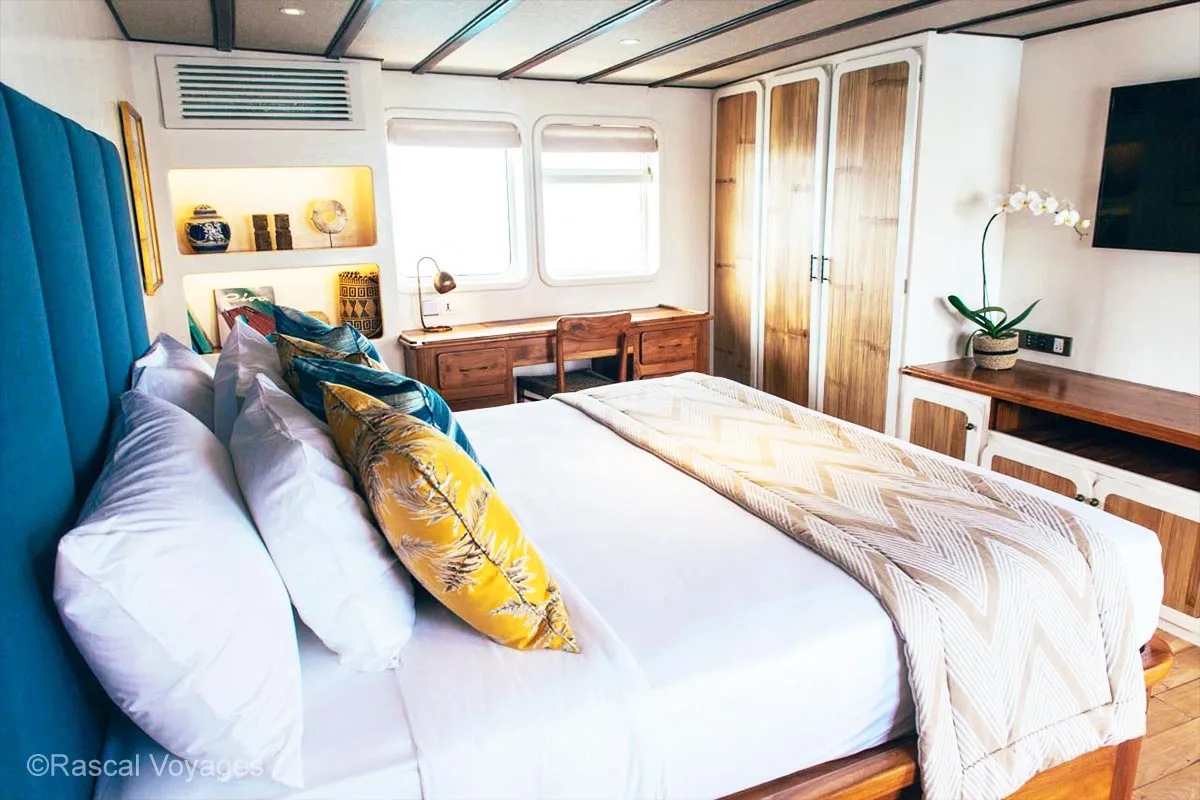 A bedroom with comfortable bed, windows, working desk, wardrobe, and a flat-screen TV. Rascal voyages. Indonesia yacht charter. Yacht charter Indonesia. Indonesia private cruise. Indonesia Luxury cruise. Bespoke Indonesia Holiday.