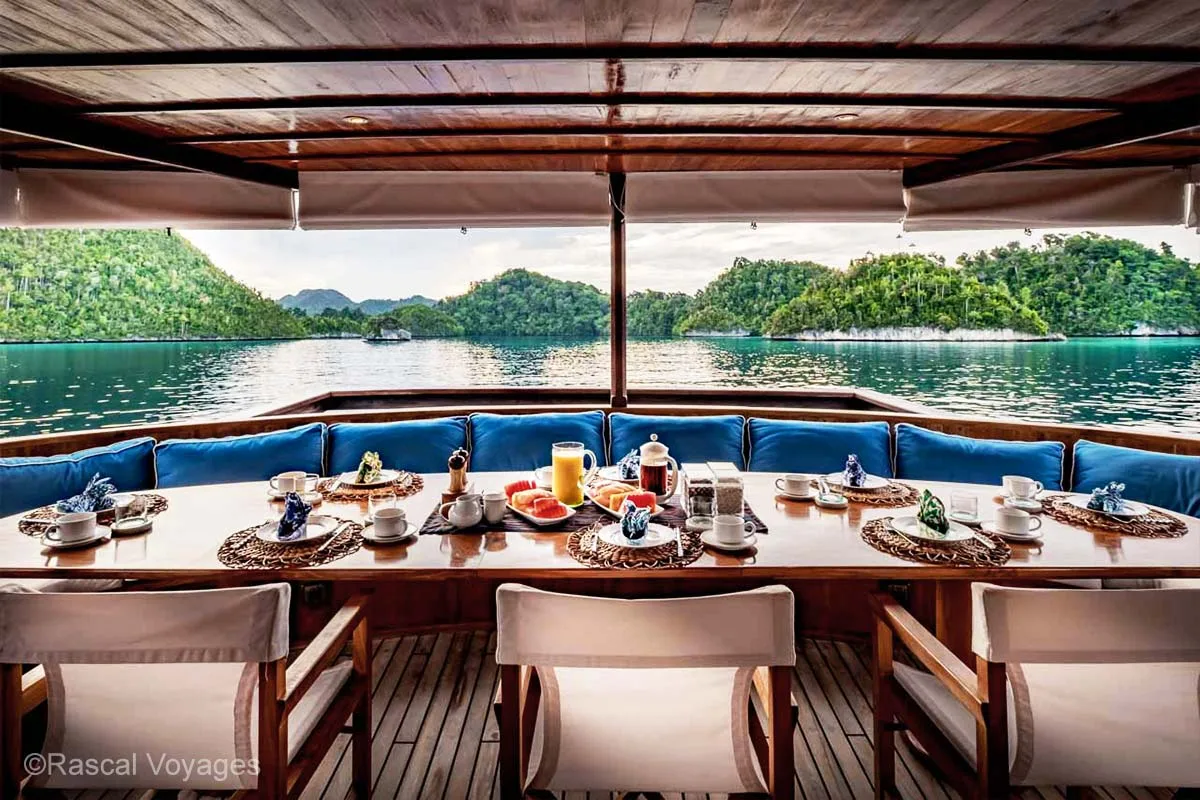 Dining table with fruits and juice on an open space with roof for shelter on a boat with views of beautiful islets in the background. Luxury sailing adventure. Rascal voyages. Indonesia yacht charter. Yacht charter Indonesia. Indonesia private cruise. Indonesia Luxury cruise. Bespoke Indonesia Holiday.