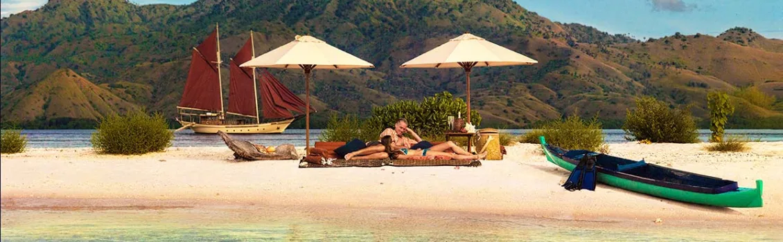 A couple relaxing on a sandbank with two white beach umbrellas behind them and green kayak in front of them. There is an island and a yellow phinisi yacht behind the sandbank with red sails. Yacht charter Indonesia. Indonesia yacht charter. Luxury cruise Indonesia. Sailing trip Indonesia. Private cruise Indonesia. Bespoke Indonesia Holiday.