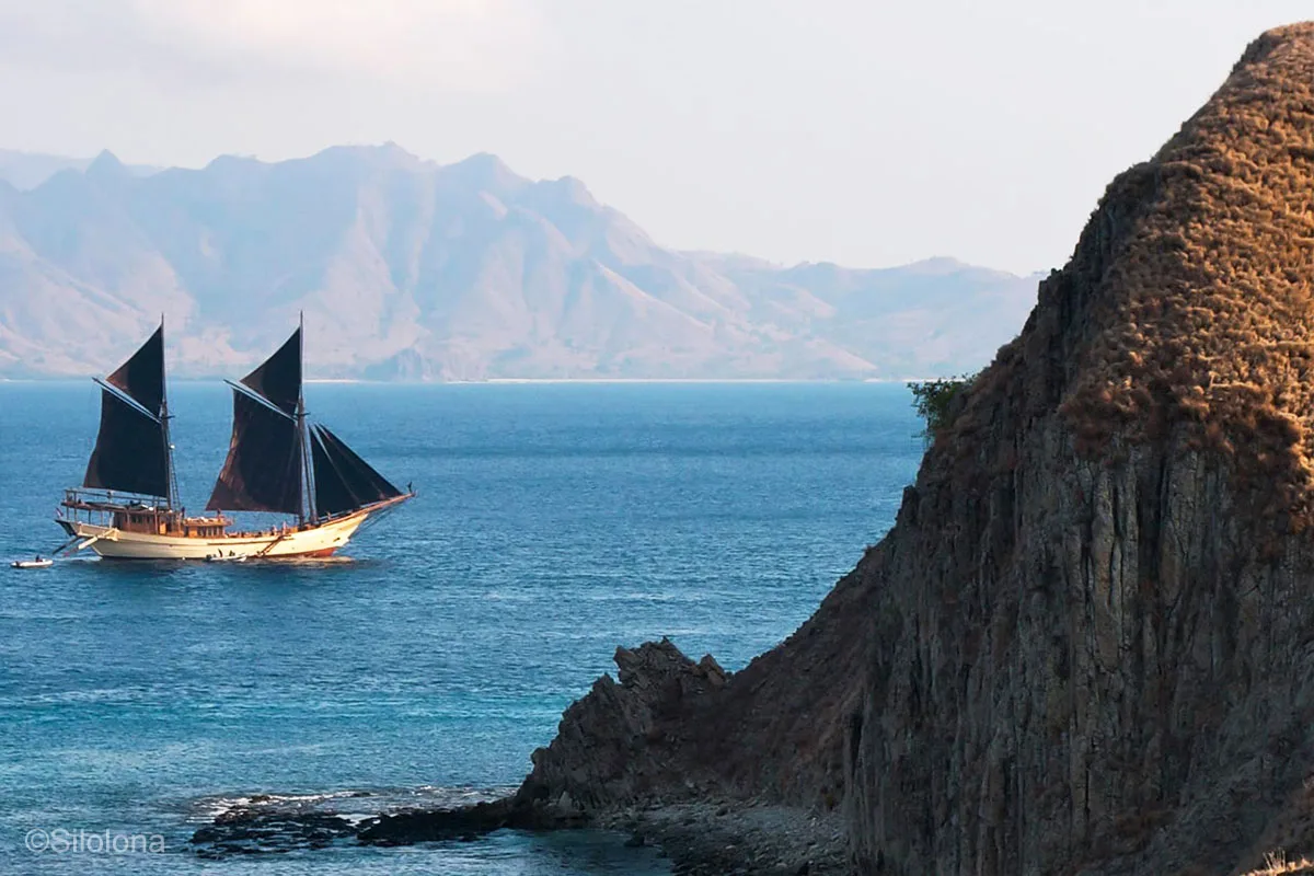 A wooden sailing boat with black sails is passing a huge rock and a big rocky island in the background. Komodo Island. Komodo National Park. Silolona. Silolona Sojourns. Indonesia sailing adventure. Yacht charter Indonesia. Komodo luxury cruises. Luxury yacht charter Komodo. Komodo liveaboard. Bespoke Indonesia Holiday.