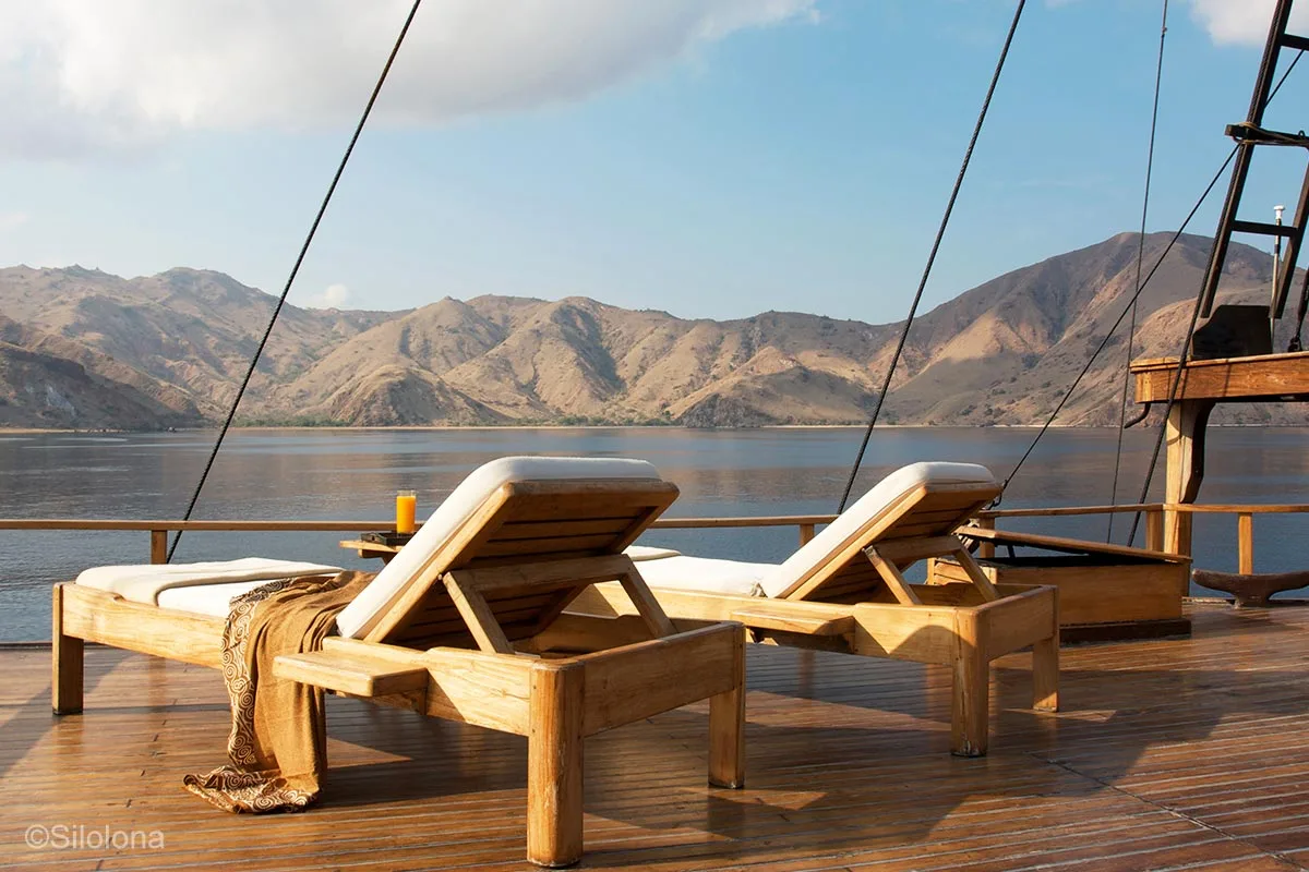 Two teak daybed loungers on an open deck area on a boat overlooking beautiful island with rugged terrain. Komodo Island. Komodo National Park. Silolona. Silolona Sojourns. Indonesia sailing adventure. Yacht charter Indonesia. Komodo luxury cruises. Luxury yacht charter Komodo. Komodo liveaboard. Bespoke Indonesia Holiday.