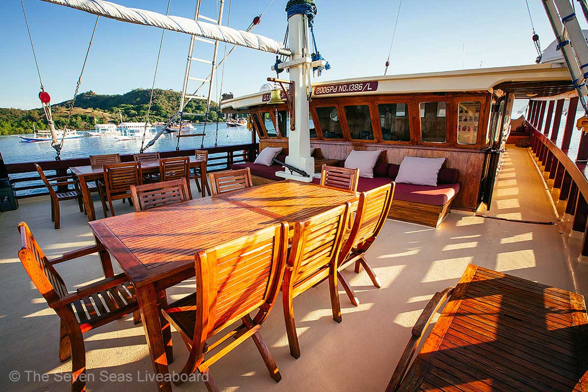 There are two sets of dining tables and a bench seat with red cushions in an open area of a wooden boat. The seven seas liveaboard. Luxury liveaboard Indonesia. Luxury liveaboard Raja Ampat. Luxury liveaboard Komodo. Diving expeditions Indonesia. Liveaboard tours Indonesia. Yacht charter Indonesia. Komodo liveaboard. Raja Ampat Liveaboard. Boat rental Indonesia. Diving trip Indonesia. Bespoke Indonesia Holiday.