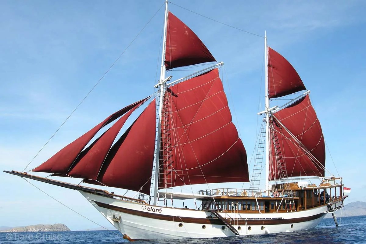 A beautiful white and natural brown wooden sailing boat with red sails. Tiare Cruise. Tiare Liveaboard. Luxury Liveaboard Indonesia. Indonesia Liveaboard. Luxury holidays Indonesia. Luxury Cruise Indonesia. Yacht charter Indonesia. Private cruise Indonesia. Sailing trip Indonesia. Yacht Rental in Indonesia. Bespoke Indonesia Holiday.