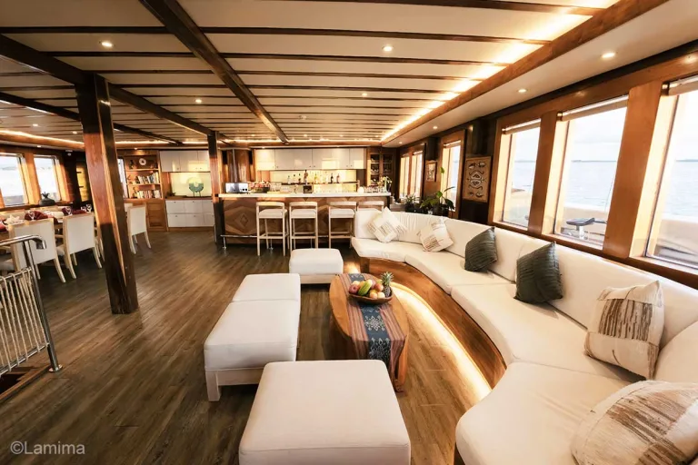There is a big white lounge on the right with fruits on the table, a bar, and dining table with white chairs. Lamima. Lamima Yacht. Lamima interior. Luxury holidays Indonesia. Luxury Cruise Indonesia. Yacht charter Indonesia. Private cruise Indonesia. Sailing trip Indonesia. Yacht Rental in Indonesia. Bespoke Indonesia Holiday.