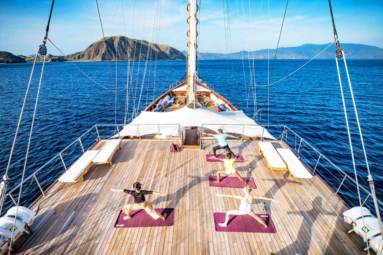 There are four people doing yoga on a wooden sailing boat while cruising toward an island. Lamima Yacht. Luxury holidays Indonesia. Luxury Cruise Indonesia. Yacht charter Indonesia. Private cruise Indonesia. Sailing trip Indonesia. Yacht Rental in Indonesia. Bespoke Indonesia Holiday.